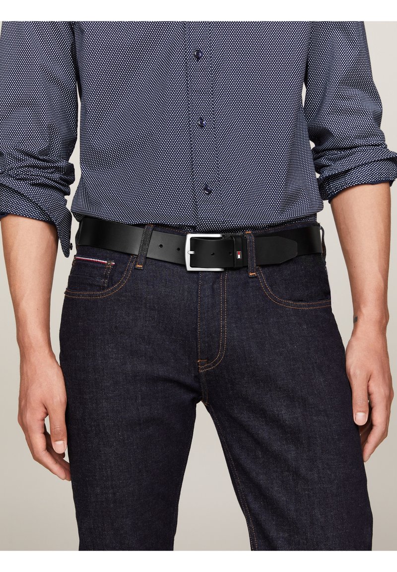 Homme portant une chemise bleu foncé à motifs rentrée dans un jean en denim foncé avec une ceinture en cuir noire et une boucle argentée.