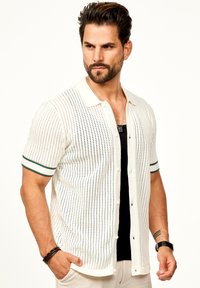 Häkelshirt mit kurzen Ärmeln in Weiß mit schwarzen Streifen, mit einer Knopfleiste, Polokragen und grün abgesetzten Ärmeln. Strukturiertes Muster über das gesamte Shirt.