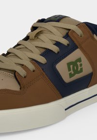 Baskets avec lacets beiges, panneaux en cuir marron et bleu marine, languette en mesh beige et logo vert de la marque DC sur le côté.