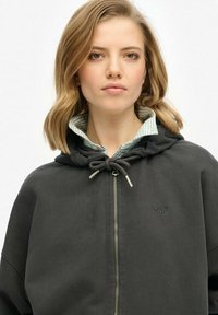 Sudadera con capucha negra y cremallera, con un detalle de cordón y un cuello a rayas verde y blanco asomándose por debajo.