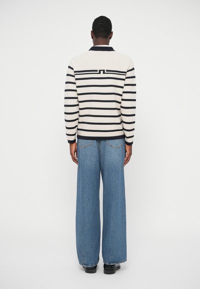 U odjeći s prugastim krem i tamnoplavim puloverom s ovratnikom, uparenim s širokim plavim trapericama, prikazano s leđa.