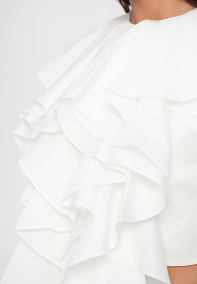 Vêtement blanc avec plusieurs volants superposés couvrant l'épaule et le haut de la poitrine, porté par une personne avec un cou visible et une partie du visage.