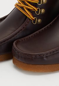 Botas de cuero marrón con punta redonda y suela de goma. Presenta cordones a rayas amarillas y rojas y ojales de tono dorado. Textura suave con detalles de costura.