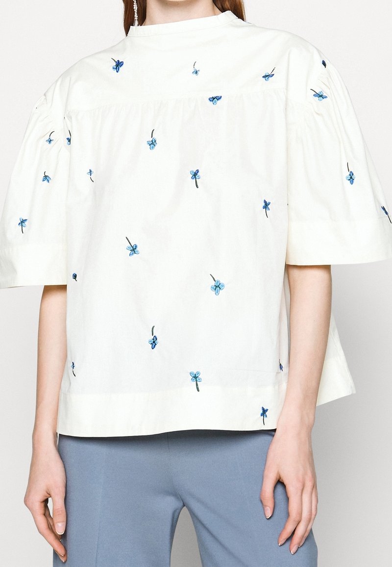 White cotton blouse with short puff sleeves, featuring blue floral embroidery throughout and a round neckline.