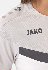 Maglietta sportiva grigio chiaro con branding JAKO, caratterizzata da un tessuto testurizzato e una striscia orizzontale grigio scuro attraversa il petto.