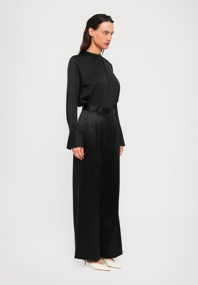 Woman with dark hair wears long-sleeve black blouse with wide cuffs, high-waisted wide black pants, and white pointed heels, standing in profile.