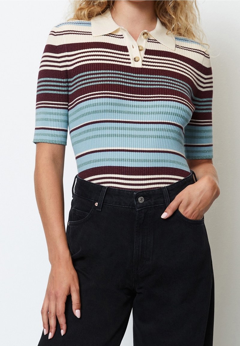 Femme portant un polo à manches courtes en maille côtelée avec des rayures horizontales bordeaux, crème et bleues, associé à un pantalon noir taille haute.