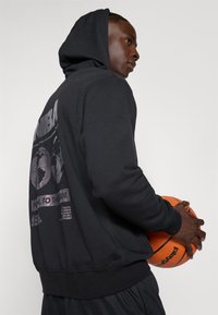 Jeune homme portant un sweat à capuche NBA noir tenant un ballon de basket orange, regardant vers la droite sur un fond uni.