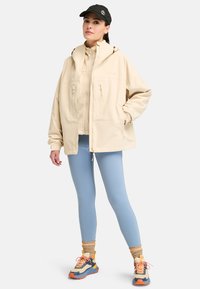 Beige väderbeständig jacka med två framfickor, tillsammans med ljusblå leggings och flerfärgade träningsskor med rosa snören.