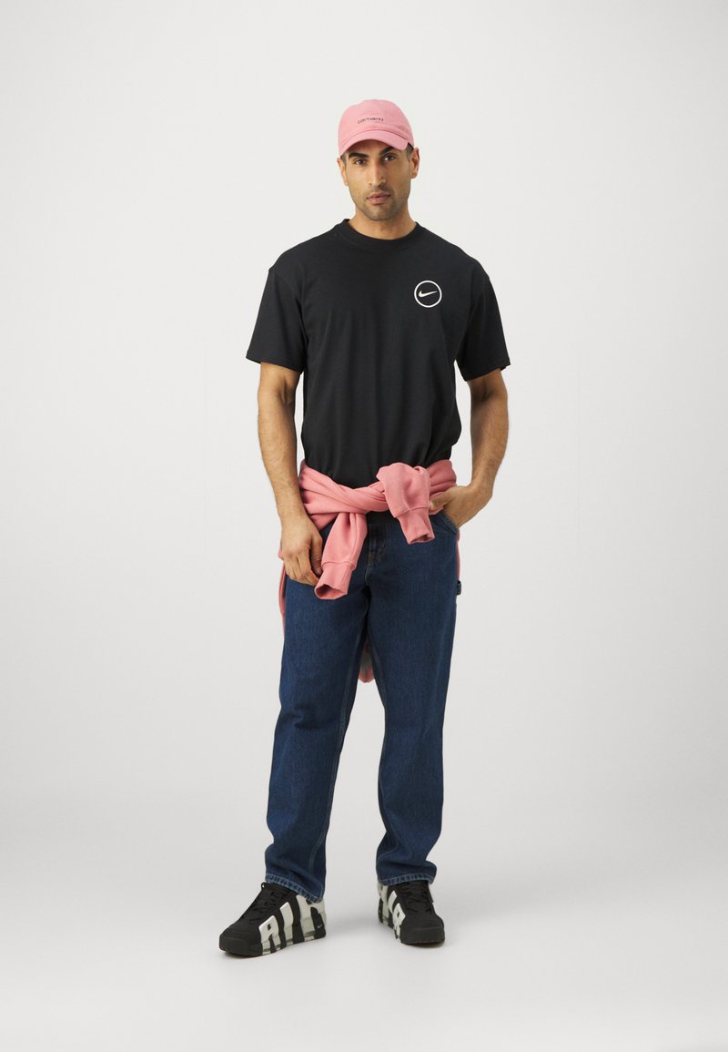 Schwarzes T-Shirt mit weißem Logo, rosa Sweatshirt um die Taille gebunden, blaue Jeans und schwarze Sneakers mit weißem Muster.