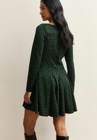 Donna con lunghi capelli scuri, che indossa un abito a maniche lunghe con stampa leopardata verde e nera e stivali neri, in posa contro un muro semplice.