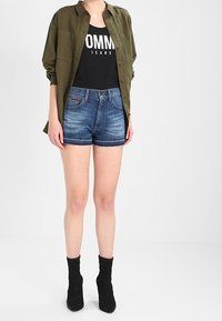 Camicia verde oliva a maniche lunghe sopra una canottiera nera con testo bianco, abbinata a pantaloni corti in denim blu e stivaletti neri alla caviglia.