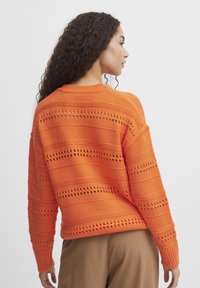 Pull en tricot orange avec un motif texturé comportant des rayures horizontales et des découpes en dentelle, encolure ronde et manches longues.