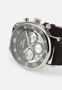 Reloj cronógrafo negro con caja de acero inoxidable plateada, números romanos, tres subdiales y correa de cuero negro. Detalles transparentes notables.
