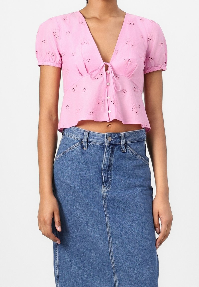 Blouse courte rose avec manches courtes bouffantes, encolure en V, fermeture à nouer sur le devant et détails floraux brodés. Associée à une jupe en jean.