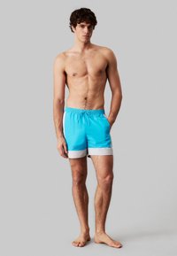 Pantaloncini da bagno azzurri con elastico in vita e coulisse. Presentano un bordo bianco nella parte inferiore. Il materiale sembra liscio e leggero.