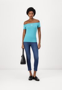 Top turquesa sin mangas, de diseño ajustado; combinado con jeans ajustados azul oscuro. Un bolso negro y mocasines completan el conjunto.