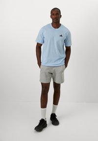 Lichtblauwe katoenen t-shirt met een zwart logo op de borst, gecombineerd met grijze sportshorts en zwarte sneakers. Model staat met de handen op de heupen.