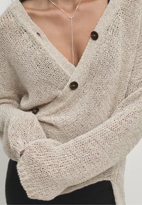 Beige gestrickte Strickjacke mit loser Maschenstruktur, die braune Knöpfe und weite Ärmel aufweist. Betonte Halslinie und strukturierter Stoff für eine entspannte Passform.
