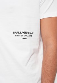 Hvit T-skjorte med svart tekst som leser "KARL LAGERFELD 21 RUE ST-GUILLAUME PARIS" på venstre bryst, båret av en person med en synlig arm.