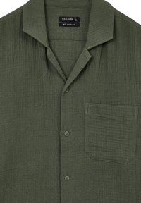 Camicia verde a maniche corte in tessuto leggero e testurizzato. Presenta un colletto, due bottoni e una tasca sul petto. Design con vestibilità rilassata.