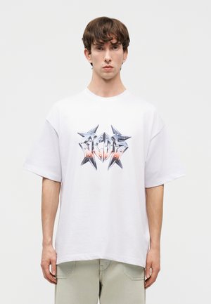 Νεαρός άνδρας με κοντά καστανά μαλλιά που φοράει μια λευκή φαρδιά μπλούζα με σχέδιο μεταλλικού αστεριού και ανοιχτόχρωμο παντελόνι.