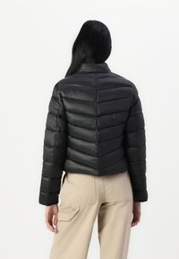 Czarna pikowana kurtka puchowa z poziomym pikowaniem, krótkim fasonem, wysokim kołnierzem i gładką fakturą. Nosi się ją na beżowych spodniach typu cargo.