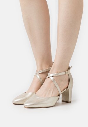 Chaussures à talons blocs dorés scintillants avec brides croisées à la cheville, fermées par de petites boucles, portées sur les pieds d’une personne sur un fond blanc uni.
