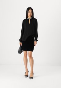 Blouse noire à manches longues avec un décolleté en forme de clé et un col orné, associée à une mini jupe noire texturée et des chaussures à talons hauts noires.