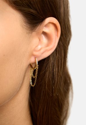 Zbliżenie na lewe ucho z małą złotą kolczykiem w kształcie koła z osadzonymi kamieniami i dwoma delikatnymi złotymi łańcuszkami zawieszonymi na nim.