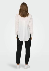 Chemise blanche à manches longues avec un ourlet arrondi et un détail de couture au dos, associée à un pantalon noir et des baskets blanches. Texture lisse, coupe décontractée.