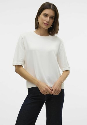 Top bianco a maniche corte realizzato in un materiale liscio. Presenta un collo a giro e maniche arrotolate, abbinato a jeans in denim scuro.