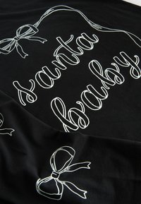 Tela negra con texto blanco que dice "santa baby", decorada con lazos de cinta ilustrados en una textura suave y lisa.