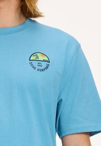 Tengezöld pamut póló rövid ujjakkal, köralakú logóval, amely pálmafákat, napot és a "SOUL SURFING" valamint a "EST. 1960" feliratokat ábrázolja.