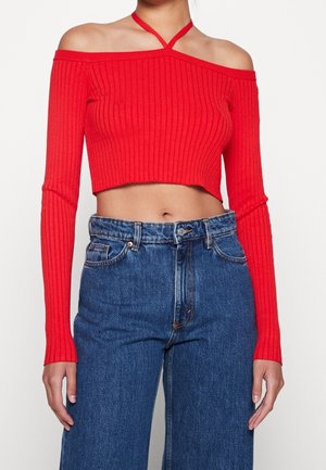 Vrouw draagt een rood off-shoulder geribd crop top met lange mouwen en hooggetailleerde blauwe spijkerbroek, staand tegen een witte achtergrond.