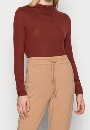 Vrouw draagt een nauwsluitende, roestkleurige top met lange mouwen, ingestopt in een beige broek met hoge taille en aantrekkoord.