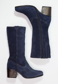 Ett par knähöga mörkblå mockastövlar med sidodragkedjor och blockklackar i metall på en vit bakgrund.