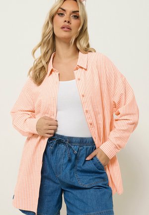 Kvinde iført løs fersken- og hvide stribede skjorte med knapper over hvid top og højtaljede blå denimshorts, poserer mod en ensfarvet baggrund.