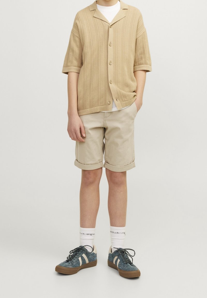 Beige gebreide korte mouwen cardigan over een wit shirt, gecombineerd met opgerolde crèmekleurige shorts en donkerblauwe sneakers met lichte accenten.
