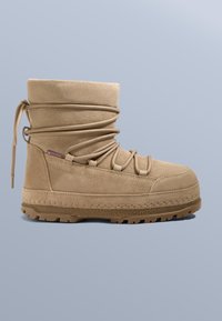 Beige mockabootie med rundad tå, tjock gummisula och korslagda snörningsdetaljer längs skaftet. Mjuk textur och enkel design.