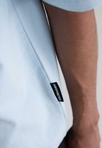 Light blue cotton t-shirt featuring a short sleeve and a black label with the word "PROHIBITED" stitched on the side seam.