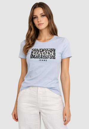 Giovane donna con lunghi capelli castani indossa una t-shirt azzurra Gaudi Jeans con logo leopardato e pantaloni bianchi contro uno sfondo semplice.