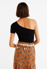 Mustassa ribattussa yhden olkapään yläosassa on kuvioitu oranssi hame, jota korostaa koristeellinen kerma-punainen vyö, jossa on tupsuja ja simpukoita.