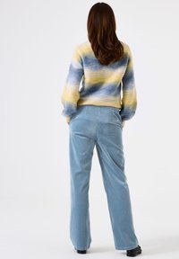 Femme aux cheveux bruns portant un pull rayé bleu, jaune et blanc et un pantalon en velours côtelé bleu clair, se tenant avec les mains dans les poches.