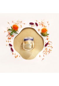 Shiseido Vital Perfection-krem i en rund, frostet glasskrukke med lokk i sølv og blått, omgitt av oransje og lilla blomsterdekorasjoner.