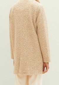 Beige ullrock med strukturerat, upphöjt prickmönster. Har långa ärmar och rak skärning, visad bakifrån, med lätt tyg under.