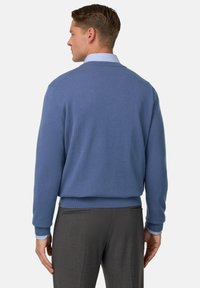 Vyrų mėlynas megztinis su apvaliu iškirpimu, smulkintais rankogaliais ir juosmeniu. Nešiojamas ant šviesiai mėlynos marškinėlių su apykakle, rodantis lygų tekstūrą.