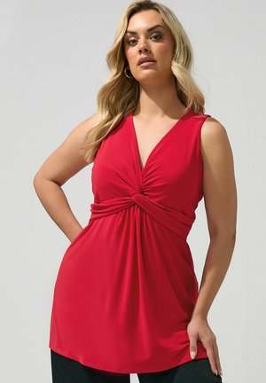 Blondynka w czerwonej sukience bez rękawów z wykręconym węzłem w talii, stojąca z jedną ręką na biodrze na jasnoszarym tle.