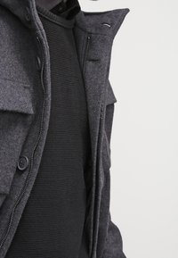 Giacca grigia in misto lana con chiusura a zip e a bottoni, quattro tasche frontali e un maglione a strati in nero testurizzato sottostante.