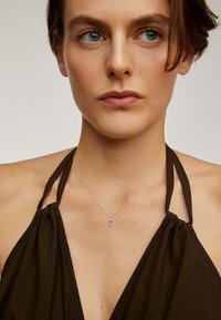 Vestido marrón de tirantes con detalle de tela fruncida, que presenta un delicado collar de plata y un pequeño colgante ovalado. Textura de piel suave.